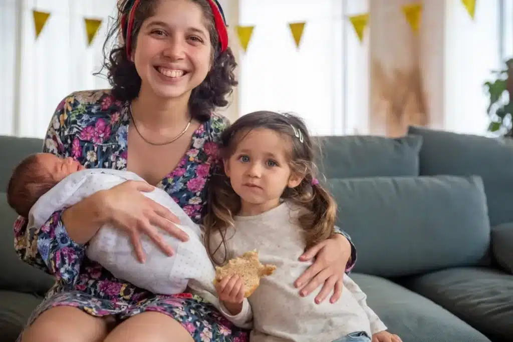 Uma jovem mãe sorridente, vestindo um vestido floral, segura um bebê recém-nascido no colo, enquanto sua filha mais velha, comendo um biscoito, senta-se ao seu lado no sofá.