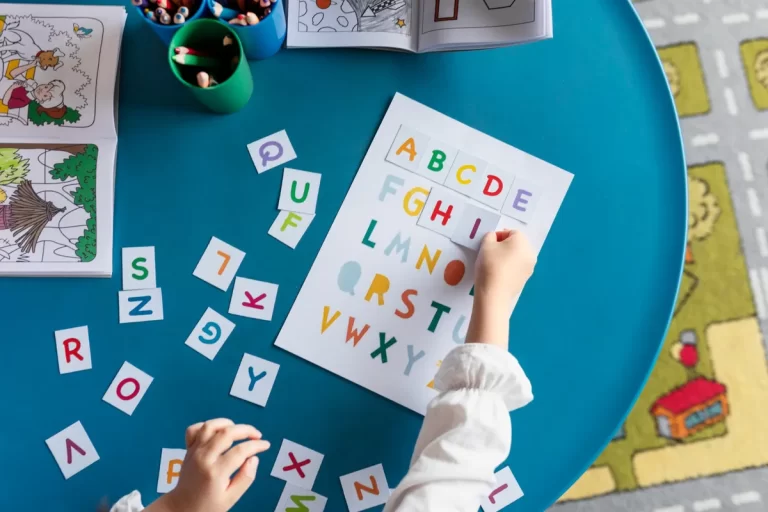Mãos de criança interagindo com letras e um cartão de alfabeto colorido em uma mesa azul, usando um método de alfabetização lúdico.