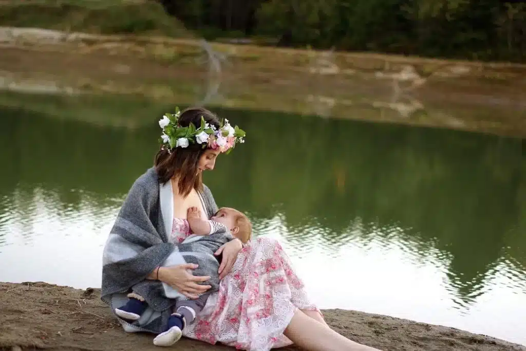 Uma mãe sentada na margem de um lago, usando uma coroa de flores e coberta por um xale, amamenta seu bebê com carinho em um ambiente natural e tranquilo