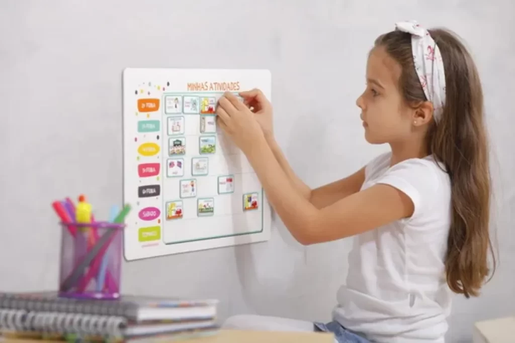 Uma menina de cabelo castanho comprido, sentada de perfil, está anexando uma figura magnética em um quadro de incentivo semanal com o título "Minhas Atividades", pendurado em uma parede cinza clara. O quadro tem espaços para os dias da semana e tarefas como "Arrume a cama" e "Escove os dentes". Há um copo com canetas e livros na mesa ao lado, sugerindo um ambiente de estudo e rotina.