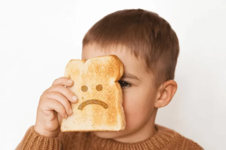 Uma criança pequena, vestindo um suéter marrom, esconde metade do rosto atrás de uma fatia de pão torrado que tem um emoticon de rosto triste (sobrancelhas, olhos e boca curvada para baixo) desenhado nela, simbolizando o desconforto ou a tristeza associada à intolerância ao glúten e à necessidade de restrição dietética.