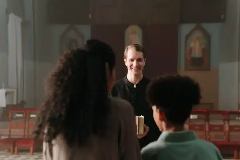 Religioso sorrindo e segurando um livro enquanto conversa com fiéis, representando o compartilhamento dos dons espirituais