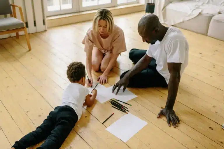 Pais sentados no chão ajudando o filho a desenhar, representando a prática da educação parental em casa