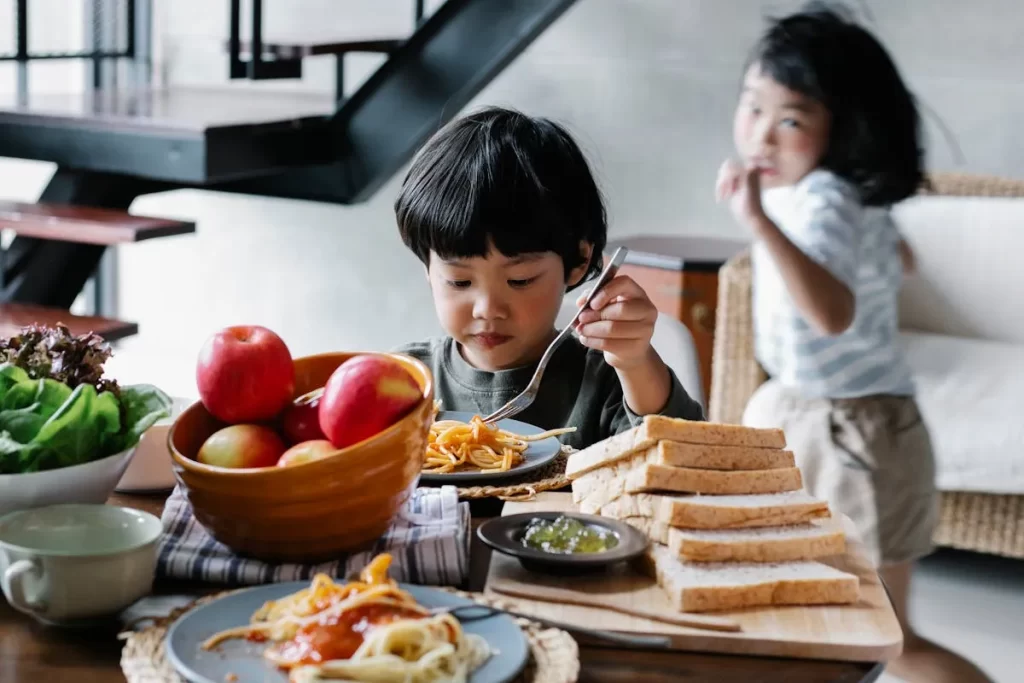 Criança comendo macarrão e frutas na mesa, representando bons hábitos alimentares na infância