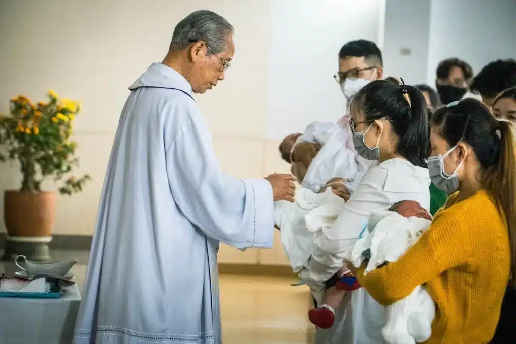 Pais segurando bebês vestidos com vestido de batismo infantil durante cerimônia religiosa com padre