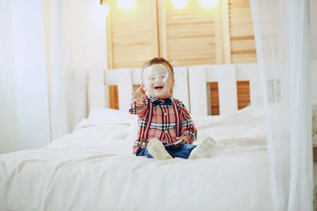 Menino bebê sorridente vestindo camisa xadrez vermelha com gravata borboleta e suspensórios, sentado no centro de um colchão amplo coberto por lençol branco texturizado.