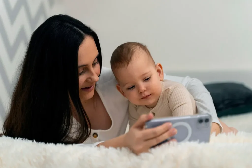 Mulher e bebê deitados de bruços em um tapete felpudo branco, olhando juntos para a tela de um celular, ilustrando o uso de monitoramento digital.