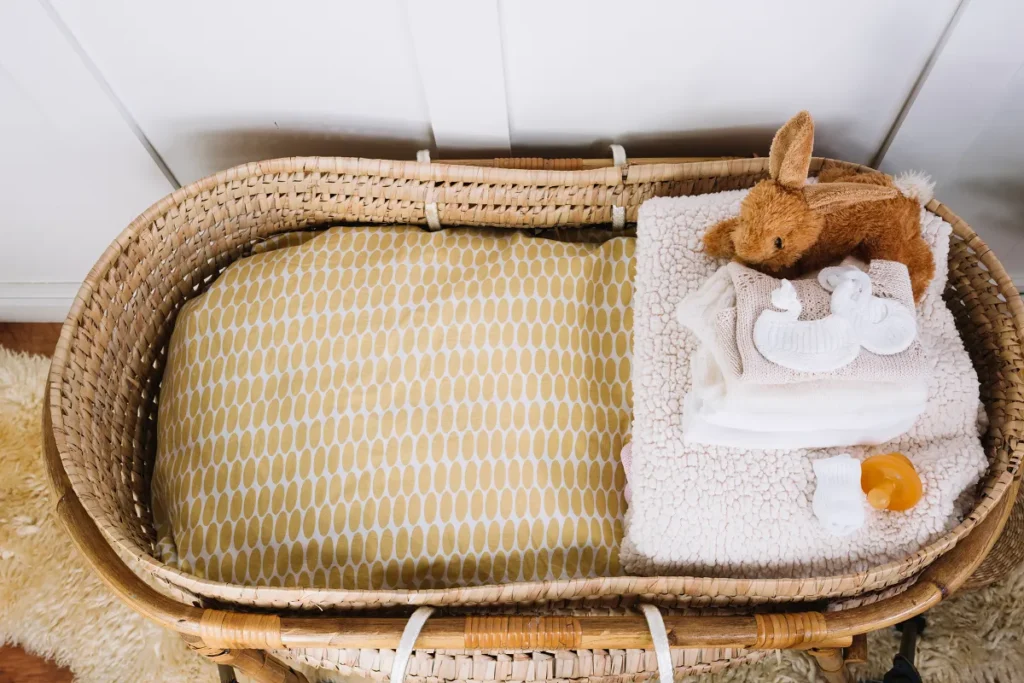 Berço portátil estilo Moisés feito de vime trançado, com enxoval amarelo e branco, roupas de bebê dobradas, chupeta e um coelho de pelúcia em cima.