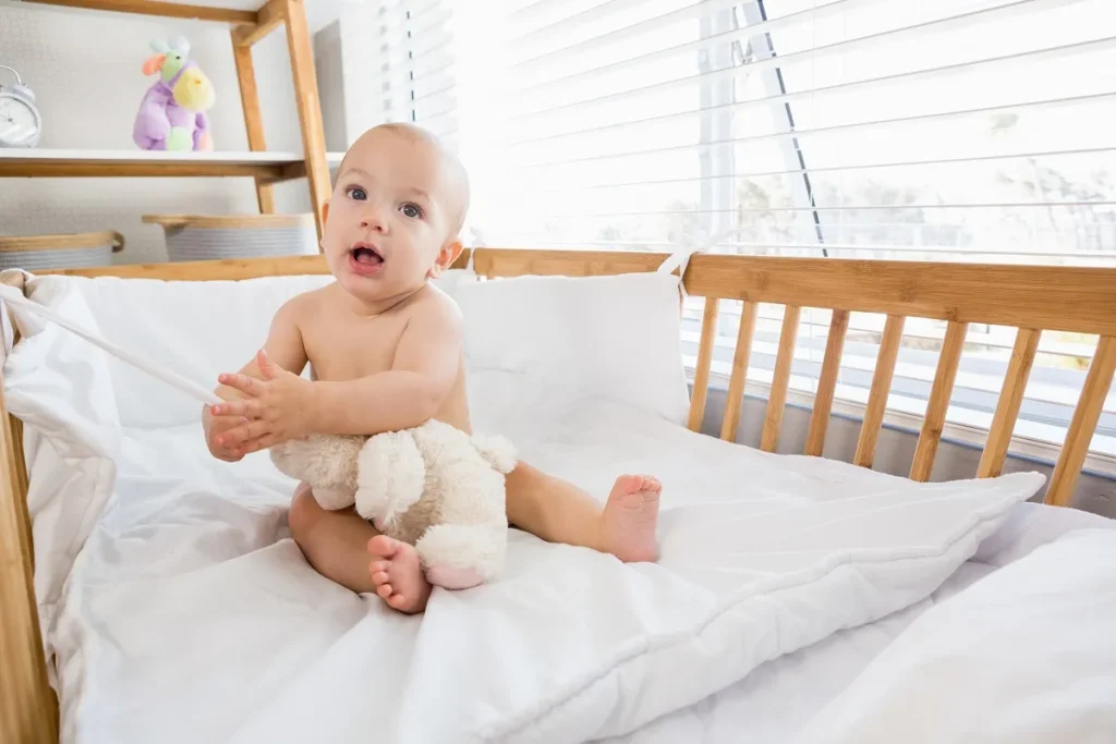 Bebê sorridente sem camisa sentado em um colchão branco e macio dentro de um berço de madeira com protetores acolchoados brancos, segurando um urso de pelúcia.