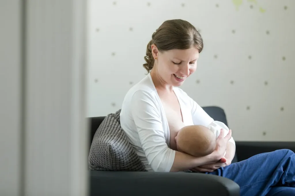 Mãe sorrindo de perfil amamentando o bebê em sofá cinza com almofada de suporte textuarda nas costas.