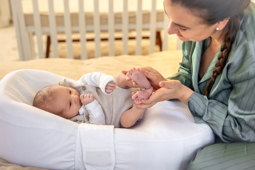 Mulher de pijama verde acetinado segurando delicadamente os pés de um bebê recém-nascido deitado em um ninho ergonômico branco sobre a cama, com um berço de madeira visível ao fundo.