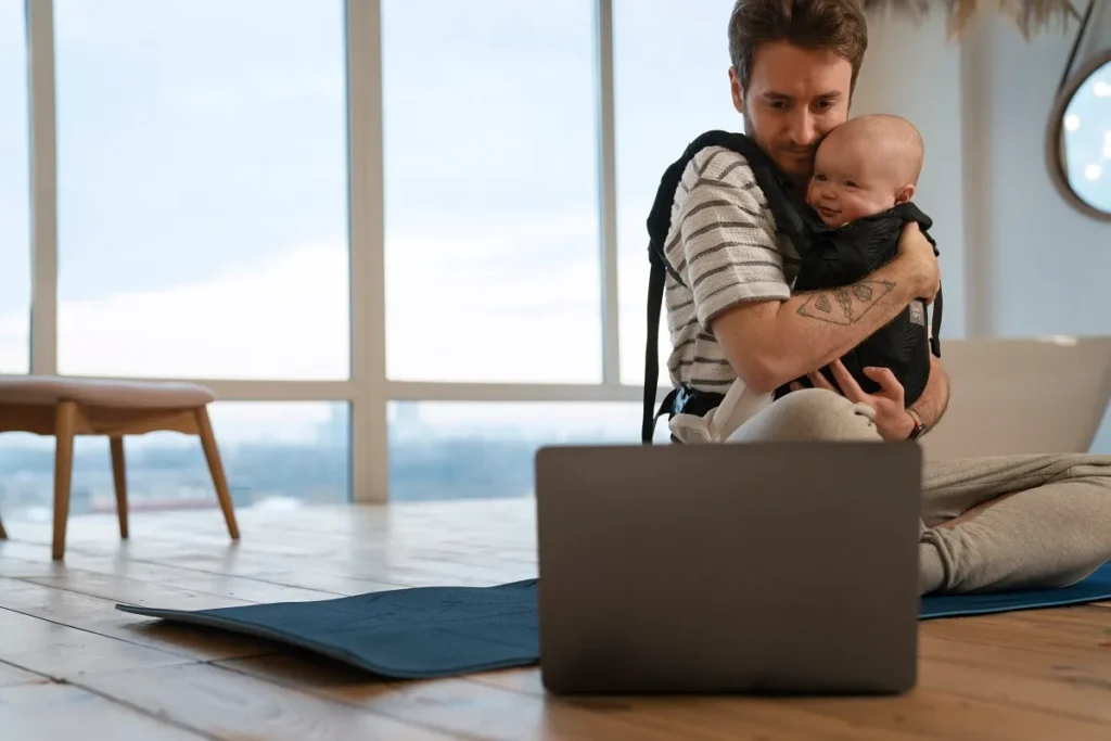 Pai sentado no chão em um tapete de ioga segurando o bebê em um canguru ergonômico enquanto observa um notebook aberto, representando o planejamento do ângulo de visão e campo óptico para monitoramento infantil.