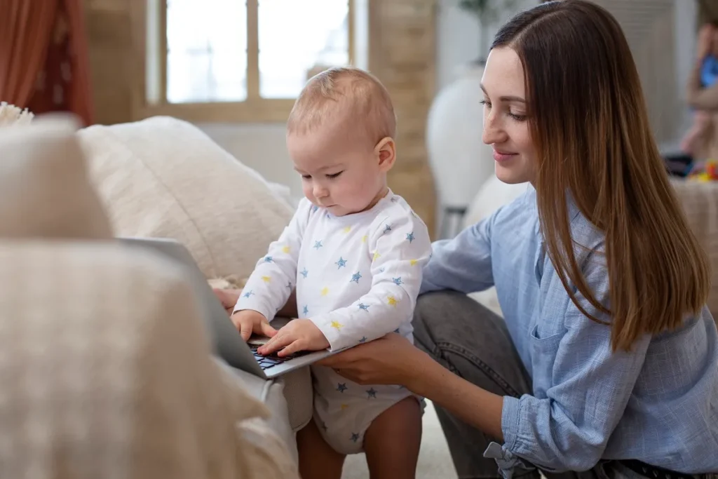 Mãe sentada no chão apoia o bebê em pé no sofá enquanto ele digita em um notebook, simbolizando a mobilidade e a escolha entre aparelhos portáteis a bateria ou conectados na tomada.