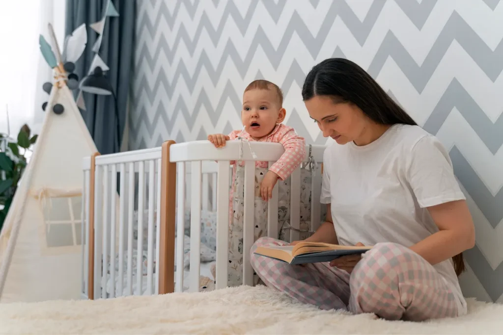 Mãe sentada no chão lendo um livro ao lado de um berço moderno de madeira branca, onde um bebê de pijama rosa está em pé apoiado na grade superior olhando para frente.