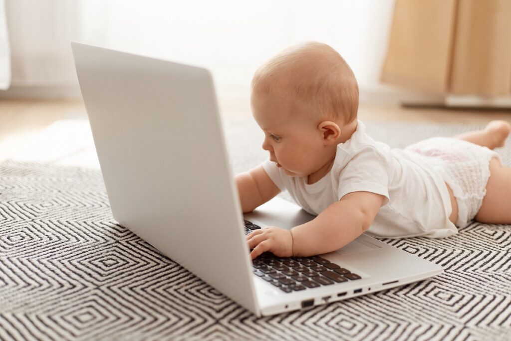 Bebê de fralda e camiseta branca deitado no tapete geométrico interagindo com um laptop prata aberto, ilustrando o conceito de tecnologia e segurança digital no monitoramento infantil.